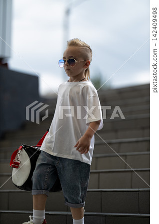 Confident young boy with a cool mohawk hairstyle wears sunglasses and casual streetwear while carrying a sports bag down modern concrete steps in an urban outdoor setting during the day. Confident young boy with a cool mohawk hairstyle wears sunglasses and casual streetwear while carrying a sports bag down modern concrete steps in an urban outdoor setting during the day. 142409498
