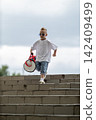 Cool young boy wearing sunglasses and casual clothes descends outdoor concrete stairs while carrying a red and white athletic duffel bag under an overcast sky. 142409499