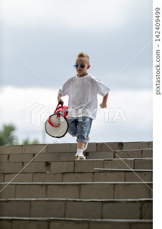 Cool young boy wearing sunglasses and casual clothes descends outdoor concrete stairs while carrying a red and white athletic duffel bag under an overcast sky. Cool young boy wearing sunglasses and casual clothes descends outdoor concrete stairs while carrying a red and white athletic duffel bag under an overcast sky. 142409499