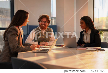 Three diverse colleagues share a relaxed team meeting around a sunlit conference table in a modern coworking office at golden hour 142411429