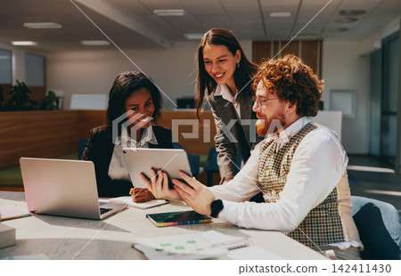 Smiling multiracial business team looks at a tablet together while collaborating in a bright modern coworking space during a workday Smiling multiracial business team looks at a tablet together while collaborating in a bright modern coworking space during a workday 142411430