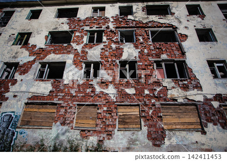 Decaying facade of an abandoned Soviet-era residential building in Kukisvumchorr, Kirovsk, Murmansk Region, Russia. Exposed red brickwork, crumbling plaster, broken windows and boarded openings show t Decaying facade of an abandoned Soviet-era residential building in Kukisvumchorr, Kirovsk, Murmansk Region, Russia. Exposed red brickwork, crumbling plaster, broken windows and boarded openings show t 142411453