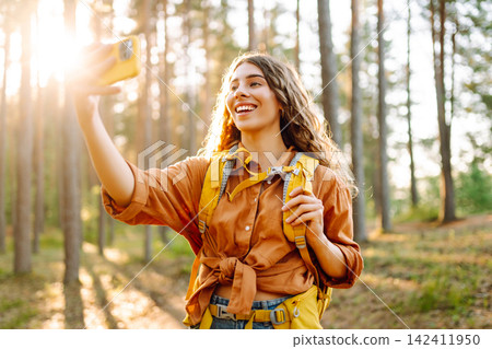 Selfie time! Young woman enjoying a sunny hike in a lush forest with a backpack on a warm day Selfie time! Young woman enjoying a sunny hike in a lush forest with a backpack on a warm day 142411950