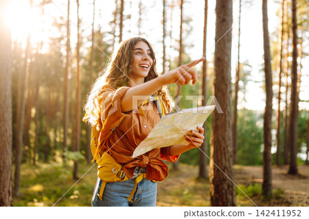 A traveler woman stands in a lush forest, examining a map with a focused expression A traveler woman stands in a lush forest, examining a map with a focused expression 142411952