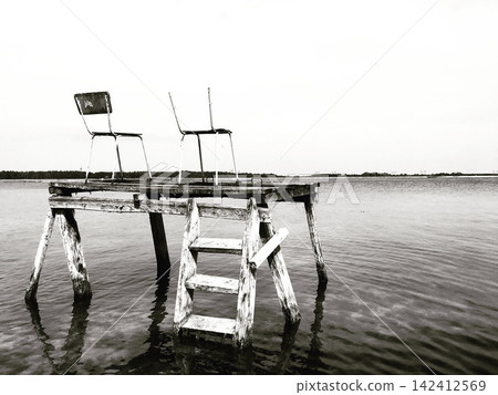 Old wooden diving pier with two chairs in calm lake, black and white monochrome Old wooden diving pier with two chairs in calm lake, black and white monochrome 142412569