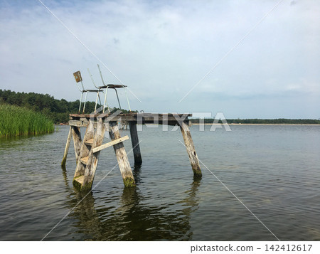 Wooden pier with ladder and chairs standing in calm lake water near green forest shore Wooden pier with ladder and chairs standing in calm lake water near green forest shore 142412617