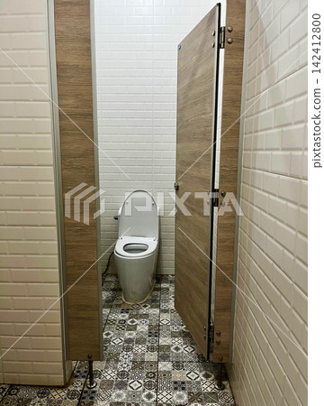 Opened wooden door reveals clean ceramic toilet bowl inside public restroom stall decorated with vintage patterned floor tiles, presenting hygienic sanitary facility Opened wooden door reveals clean ceramic toilet bowl inside public restroom stall decorated with vintage patterned floor tiles, presenting hygienic sanitary facility 142412800