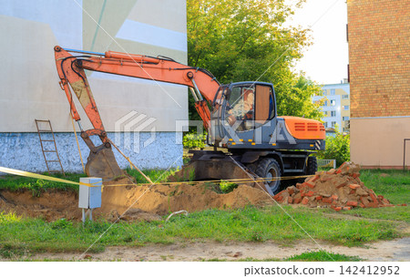 Wheeled Digger Machine Excavating Ground for Heating Utility Pipeline by Residential Block Wheeled Digger Machine Excavating Ground for Heating Utility Pipeline by Residential Block 142412952