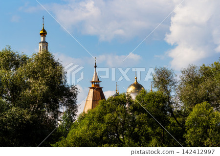 Golden domes of an Orthodox monastery rise above lush green trees under a bright blue sky, showcasing traditional Russian religious architecture and heritage in a serene summer landscape, Russia Golden domes of an Orthodox monastery rise above lush green trees under a bright blue sky, showcasing traditional Russian religious architecture and heritage in a serene summer landscape, Russia 142412997