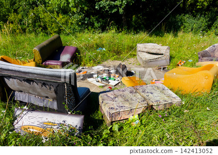 Abandoned mismatched vintage furniture scattered in a grassy meadow forming a derelict outdoor lounge area with litter and debris left behind in a forest clearing Abandoned mismatched vintage furniture scattered in a grassy meadow forming a derelict outdoor lounge area with litter and debris left behind in a forest clearing 142413052