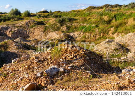 Rugged terrain featuring mounds of loose earth, rocky soil, and scattered stones, highlighting a raw industrial landscape reclaimed by wild grasses and shrubs under a bright sunny sky Rugged terrain featuring mounds of loose earth, rocky soil, and scattered stones, highlighting a raw industrial landscape reclaimed by wild grasses and shrubs under a bright sunny sky 142413061