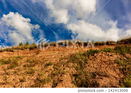 Rugged clay cliff side featuring uneven earthy textures and sparse wild vegetation under a vibrant blue sky with fluffy white clouds, capturing a raw geological landscape perspective Rugged clay cliff side featuring uneven earthy textures and sparse wild vegetation under a vibrant blue sky with fluffy white clouds, capturing a raw geological landscape perspective 142413098