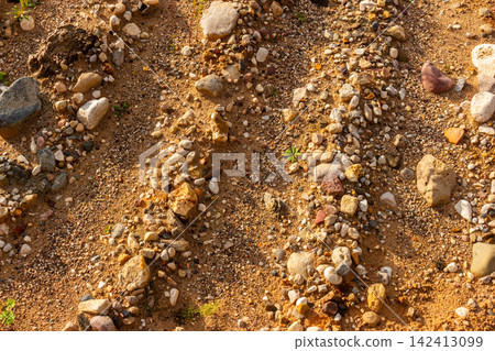 Textured surface of an abandoned gravel quarry featuring scattered pebbles, rough stones, and golden sand under warm sunlight in a detailed top-down view Textured surface of an abandoned gravel quarry featuring scattered pebbles, rough stones, and golden sand under warm sunlight in a detailed top-down view 142413099