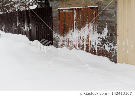 Rustic wooden garage doors weathered by time and harsh winter, covered with fresh white snow drifts beside a dark corrugated metal fence in a quiet village setting at daytime Rustic wooden garage doors weathered by time and harsh winter, covered with fresh white snow drifts beside a dark corrugated metal fence in a quiet village setting at daytime 142413107