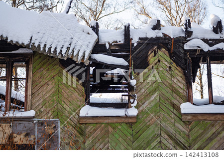 Burnt wooden house ruins covered with fresh white snow, charred timber frame and empty window frames in a winter forest, desolate structural decay after a fire incident Burnt wooden house ruins covered with fresh white snow, charred timber frame and empty window frames in a winter forest, desolate structural decay after a fire incident 142413108