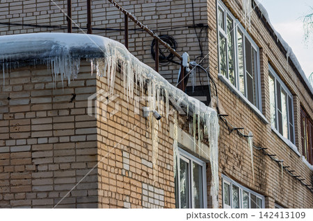 Sharp icicles hang dangerously from the edge of a brick building roof under a layer of frozen winter snow, highlighting the cold urban landscape during a freezing weather season Sharp icicles hang dangerously from the edge of a brick building roof under a layer of frozen winter snow, highlighting the cold urban landscape during a freezing weather season 142413109