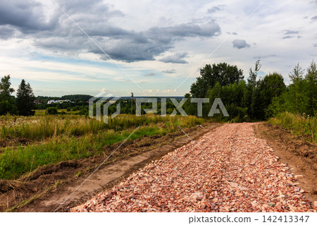 Rough rural road under construction featuring a thick layer of crushed red bricks, stretching through a picturesque countryside landscape with lush green fields and moody summer skies Rough rural road under construction featuring a thick layer of crushed red bricks, stretching through a picturesque countryside landscape with lush green fields and moody summer skies 142413347