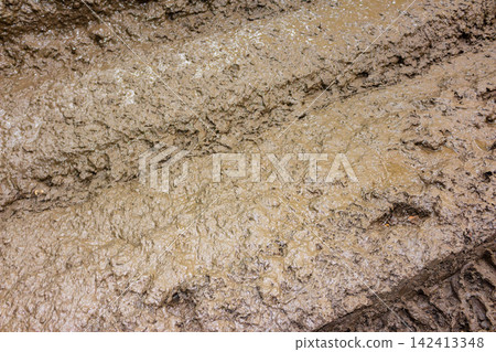 Detailed close-up of thick wet brown mud with deep tire tracks and rough soil texture covering a rural path, perfect for off-road adventure or environmental background concepts Detailed close-up of thick wet brown mud with deep tire tracks and rough soil texture covering a rural path, perfect for off-road adventure or environmental background concepts 142413348