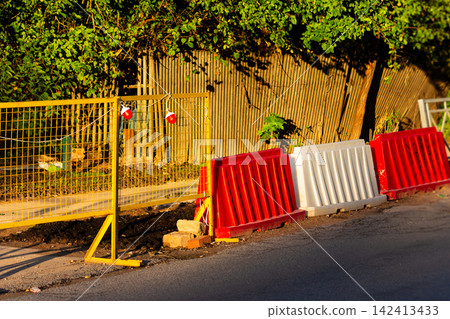 Yellow metal fencing with warning lights and plastic road barriers marking a construction zone along the street during golden hour Yellow metal fencing with warning lights and plastic road barriers marking a construction zone along the street during golden hour 142413433