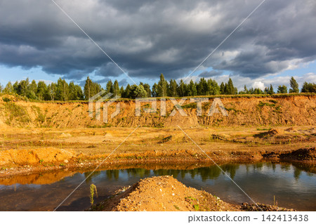 Abandoned sand quarry with orange clay banks, small pond with reflection and lush green trees against a dramatic moody stormy sky landscape in an open excavation site 142413438