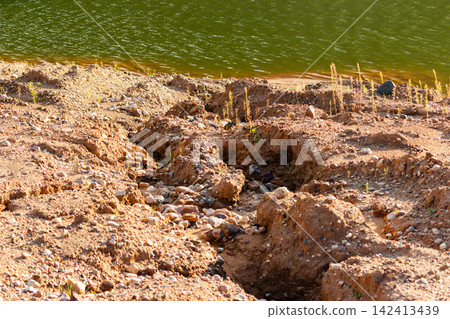 Severe soil erosion along a sandy quarry bank showing deep furrows and washed-out terrain near a water reservoir created by natural rainfall runoff and geological landscape instability 142413439
