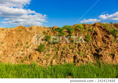 Large pile of raw soil and earth mounds in a green field, showing traces of heavy machinery excavation work under a vibrant blue sky with soft white clouds Large pile of raw soil and earth mounds in a green field, showing traces of heavy machinery excavation work under a vibrant blue sky with soft white clouds 142413488