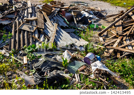 Messy pile of rural waste with old wooden boards, discarded corrugated slate sheets, plastic bags, and construction debris dumped illegally on green grass 142413495