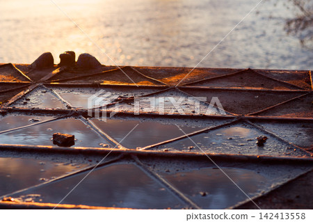 Close-up of a weathered metal pontoon dock with geometric steel frames and puddles, capturing the soft warm glow of a setting sun reflecting off the tranquil river surface at twilight Close-up of a weathered metal pontoon dock with geometric steel frames and puddles, capturing the soft warm glow of a setting sun reflecting off the tranquil river surface at twilight 142413558