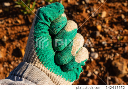 Worker's hand in a green protective glove clenched into a tight fist close-up, symbolizing manual labor, agriculture and gardening against blurry soil background Worker's hand in a green protective glove clenched into a tight fist close-up, symbolizing manual labor, agriculture and gardening against blurry soil background 142413652