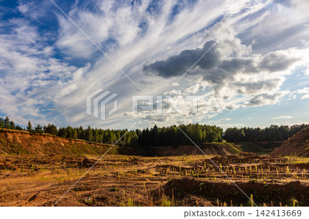 Vast sandy quarry with rugged textured ground and scattered greenery under a dramatic sky with wispy clouds, capturing the raw beauty of an industrial landscape at golden hour 142413669