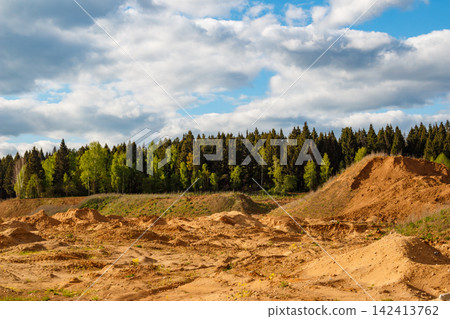 Expansive view of a textured sand quarry featuring piles of loose soil under a dramatic blue sky with fluffy white clouds, bordering a dense coniferous and deciduous forest backdrop 142413762