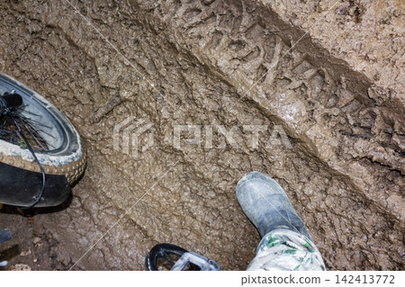 Detailed close-up of a deep tire track imprinted in thick wet mud with a bicycle wheel and a rubber boot visible in the frame, capturing a rugged off-road adventure scene Detailed close-up of a deep tire track imprinted in thick wet mud with a bicycle wheel and a rubber boot visible in the frame, capturing a rugged off-road adventure scene 142413772