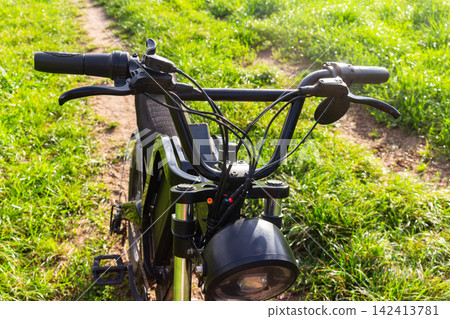 Vew of a modern electric bike handlebar, sleek black metal controls, brake levers, and headlight detail set against a vibrant sunlit grassy meadow country trail Vew of a modern electric bike handlebar, sleek black metal controls, brake levers, and headlight detail set against a vibrant sunlit grassy meadow country trail 142413781