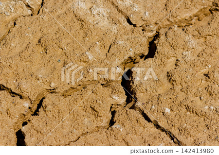 Top view of a sun-baked mud surface with deep cracks and uneven texture, showing dry earthy ground patterns caused by intense summer heat and dehydration of the land Top view of a sun-baked mud surface with deep cracks and uneven texture, showing dry earthy ground patterns caused by intense summer heat and dehydration of the land 142413980