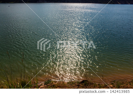 Sparkling blue water surface reflecting bright sunbeams, creating a shimmering effect on the rippling lake expanse, observed from a wild grassy shore during a clear sunny day Sparkling blue water surface reflecting bright sunbeams, creating a shimmering effect on the rippling lake expanse, observed from a wild grassy shore during a clear sunny day 142413985