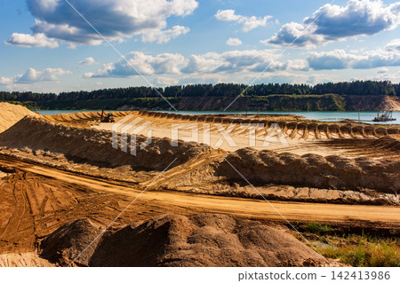 Active industrial sand quarry with massive golden dunes, heavy extraction machinery, and turquoise water pit under a bright cloudy sky Active industrial sand quarry with massive golden dunes, heavy extraction machinery, and turquoise water pit under a bright cloudy sky 142413986