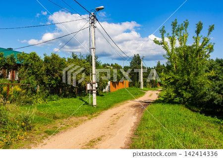 Concrete utility pole standing tall on a rustic dirt road in a quiet countryside village under a bright sunny summer sky with lush greenery and rural houses around Concrete utility pole standing tall on a rustic dirt road in a quiet countryside village under a bright sunny summer sky with lush greenery and rural houses around 142414336