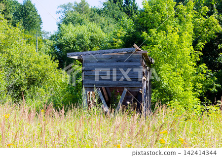 Weathered wooden shed standing alone in a wild overgrown meadow, rustic shack with cracked boards surrounded by lush green summer forest, derelict timber structure in rural countryside Weathered wooden shed standing alone in a wild overgrown meadow, rustic shack with cracked boards surrounded by lush green summer forest, derelict timber structure in rural countryside 142414344