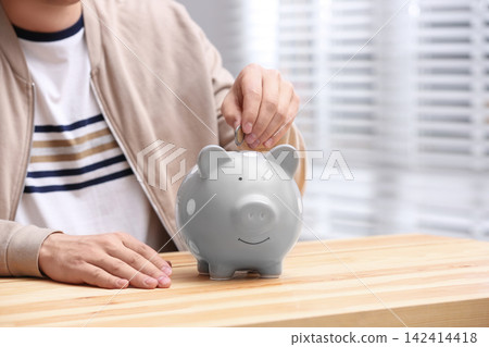 Man putting coin into piggy bank at wooden table, closeup Man putting coin into piggy bank at wooden table, closeup 142414418