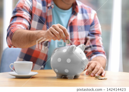 Man putting money into piggy bank at table, closeup Man putting money into piggy bank at table, closeup 142414423