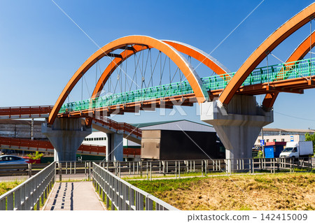 Modern orange arch pedestrian bridge spanning over a bustling highway with moving trucks and cars on a bright sunny day against a clear blue sky, architectural urban structure 142415009