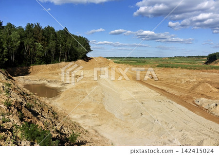 Vast industrial sand quarry with exposed earth layers and mud puddles bordered by a lush green forest under a brilliant blue sky with soft white clouds on a sunny day Vast industrial sand quarry with exposed earth layers and mud puddles bordered by a lush green forest under a brilliant blue sky with soft white clouds on a sunny day 142415024