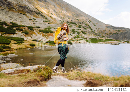 Hiking near a lake in mountain area with a woman wearing a yellow jacket and backpack during daytime Hiking near a lake in mountain area with a woman wearing a yellow jacket and backpack during daytime 142415181
