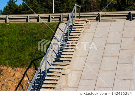 Concrete pedestrian staircase with metal railings leading up a steep highway embankment side, bright sunny day with natural light and distinct shadows on the slope Concrete pedestrian staircase with metal railings leading up a steep highway embankment side, bright sunny day with natural light and distinct shadows on the slope 142415294