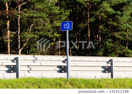 Blue highway kilometer marker sign displaying number 113 standing near metal safety guardrail against a dense green pine forest background in sunny summer daylight Blue highway kilometer marker sign displaying number 113 standing near metal safety guardrail against a dense green pine forest background in sunny summer daylight 142415296