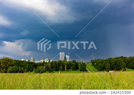 Dark ominous storm clouds gather over a vibrant green meadow and a lush forest with modern city buildings rising on the horizon under a dramatic moody sky before the heavy rain Dark ominous storm clouds gather over a vibrant green meadow and a lush forest with modern city buildings rising on the horizon under a dramatic moody sky before the heavy rain 142415300