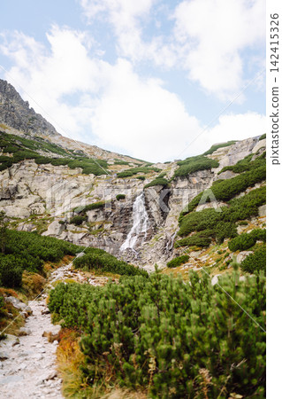Waterfall flows down rocky slope in mountain area during daytime under partly cloudy sky 142415326