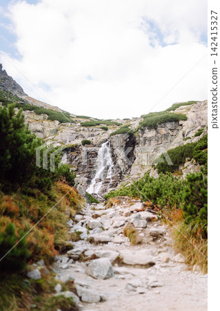 Waterfall flows down rocky slope in mountain area during daytime under partly cloudy sky 142415327