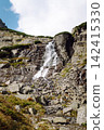 Waterfall flows down rocky slope in mountain area during daytime under partly cloudy sky 142415330