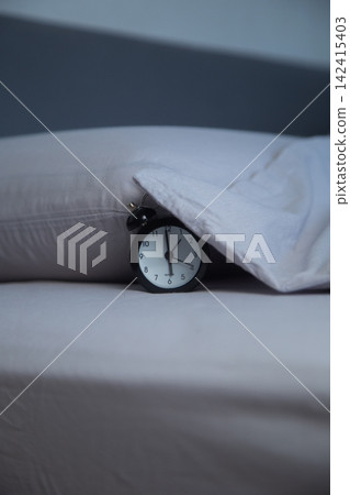 Vertical photo of a black alarm clock lying on the bed next to a pillow, partially covered with the edge of a gray blanket on a gray background. The concept of getting up in the morning, sleeping Vertical photo of a black alarm clock lying on the bed next to a pillow, partially covered with the edge of a gray blanket on a gray background. The concept of getting up in the morning, sleeping 142415403